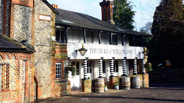 A traditional English pub, The Duke of Wellington Surrey features white walls, black trim, brick and stone details, large lanterns, and wooden barrels by the entrance on a sunny day.