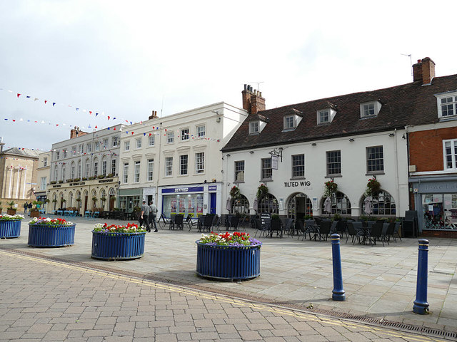 A town square with flower tubs, outdoor seating, and historic buildings, including one with a sign reading TILTED WIG. Strings of bunting hang overhead on a cloudy day near The Rose & Crown Warwickshire.