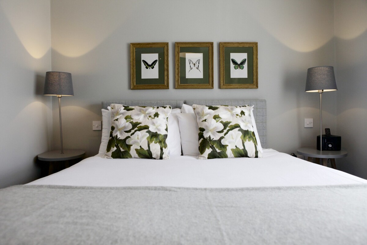 A neatly made bed with white and floral pillows, flanked by bedside tables and grey lamps. Above the bed, three framed butterfly prints decorate the light-coloured wall, evoking the charm of The Rose & Crown Warwickshire.