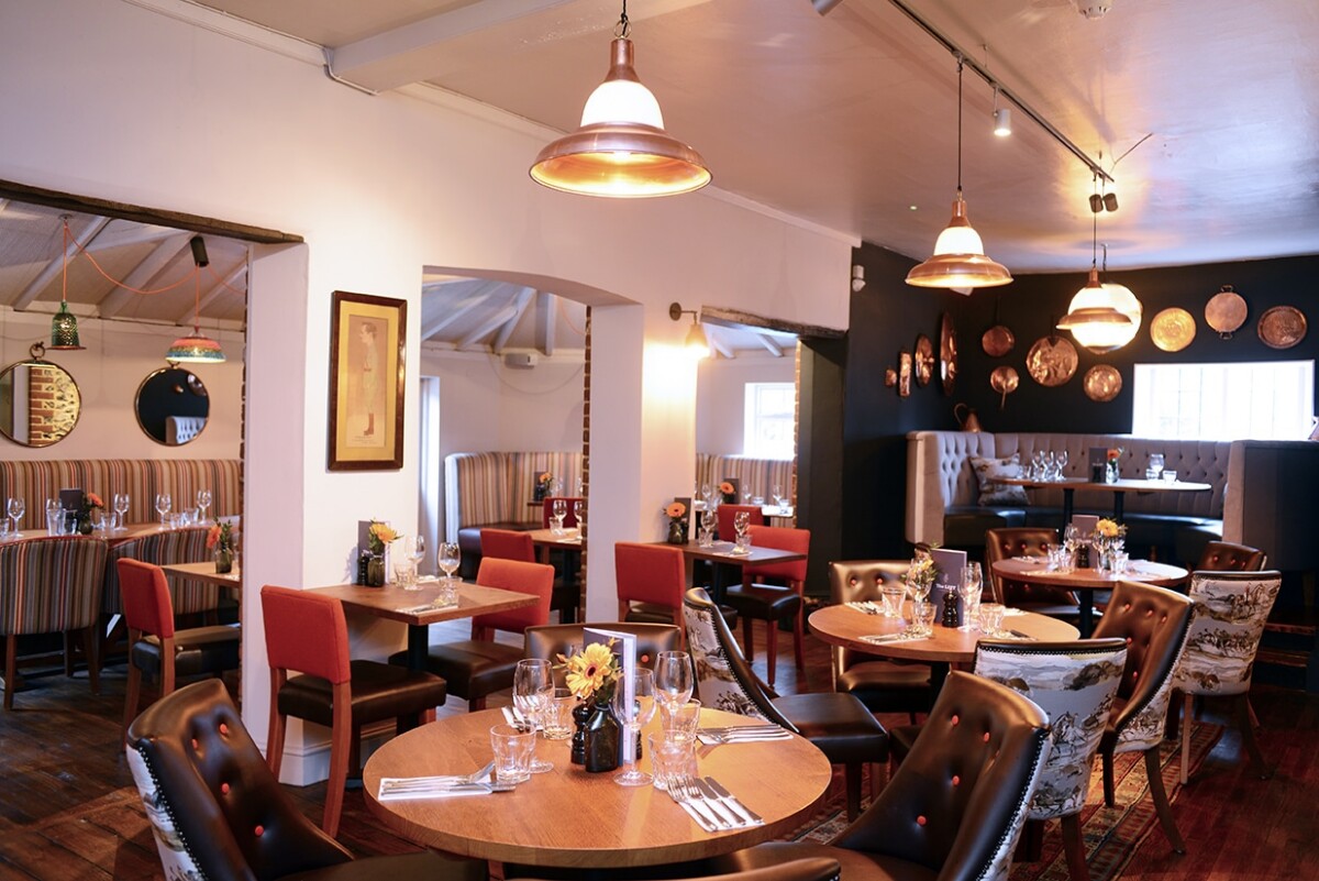 A cosy restaurant interior with round wooden tables set for dining, cushioned booths, and red chairs. Warm pendant lights hang from the ceiling, and copper pans decorate a dark feature wall.