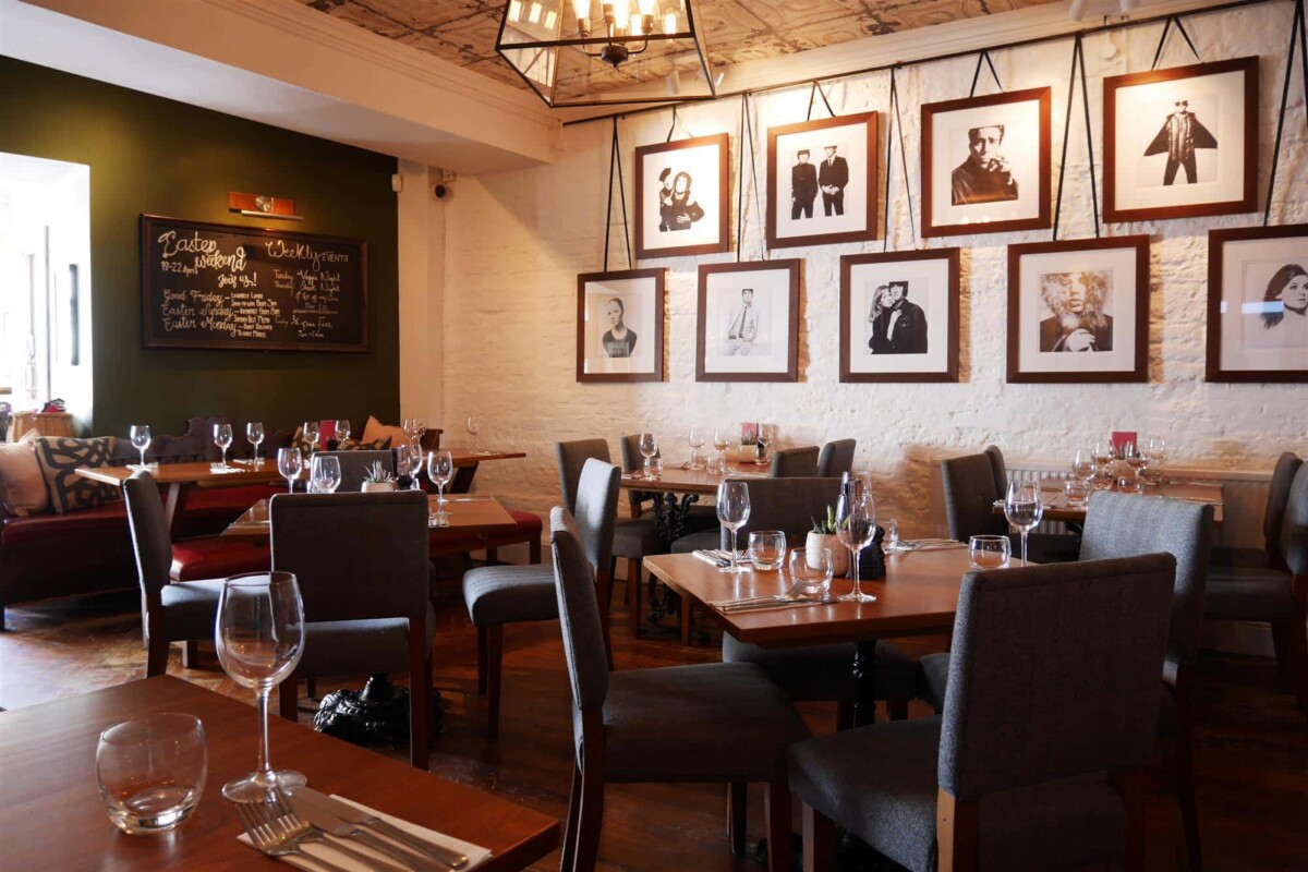 A stylish restaurant interior with white brick walls adorned with framed black and white photos. Dining tables are set with wine glasses and cutlery.