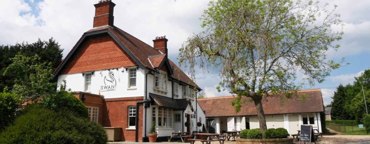 A traditional English pub called The Swan with red brick and white walls, outdoor seating, a tree in the foreground, and a blue sky with scattered clouds.