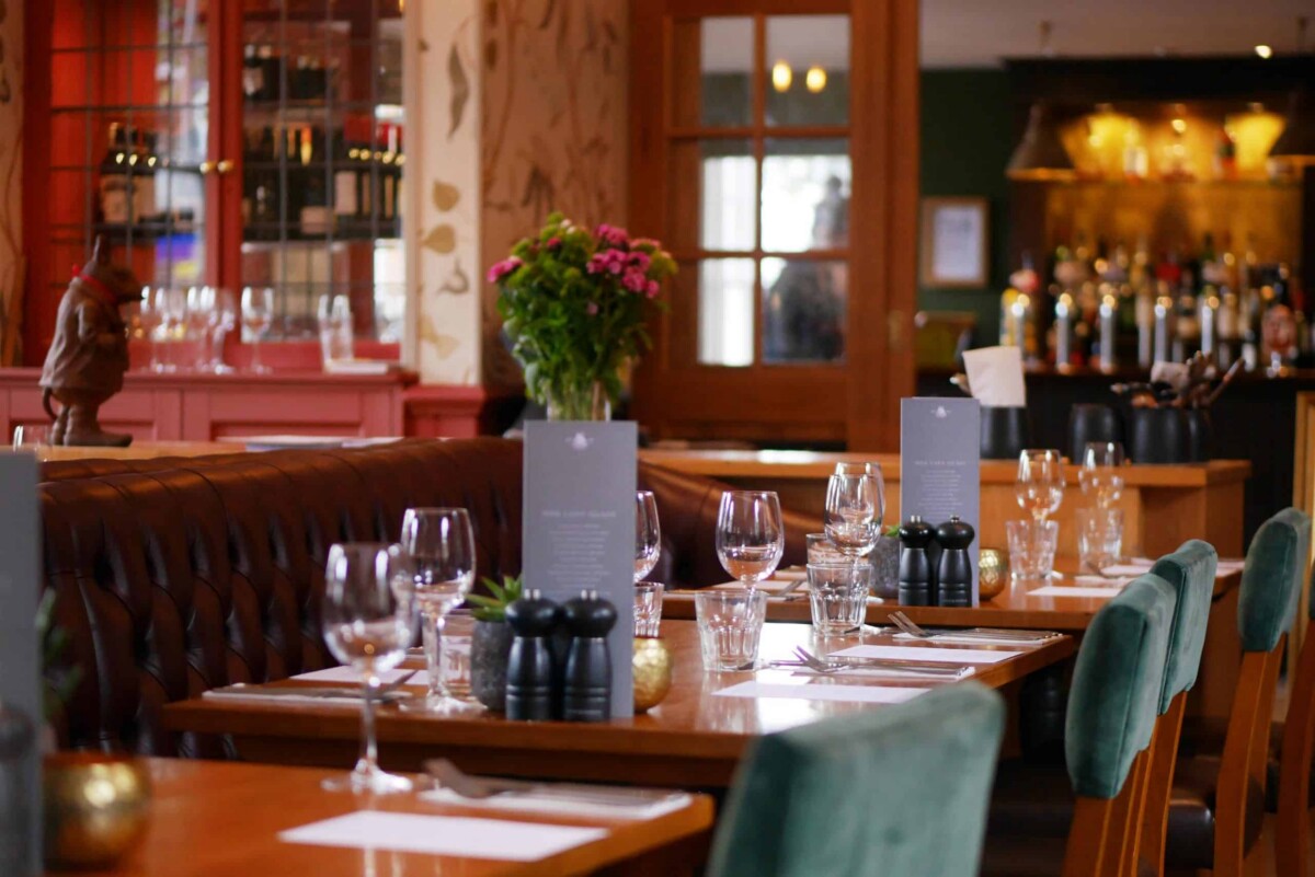 A cosy restaurant interior with tables set for dining, featuring wine glasses, menus, cutlery, and serviettes. There are green chairs, flowers as centrepieces, and a bar with bottles in the background.