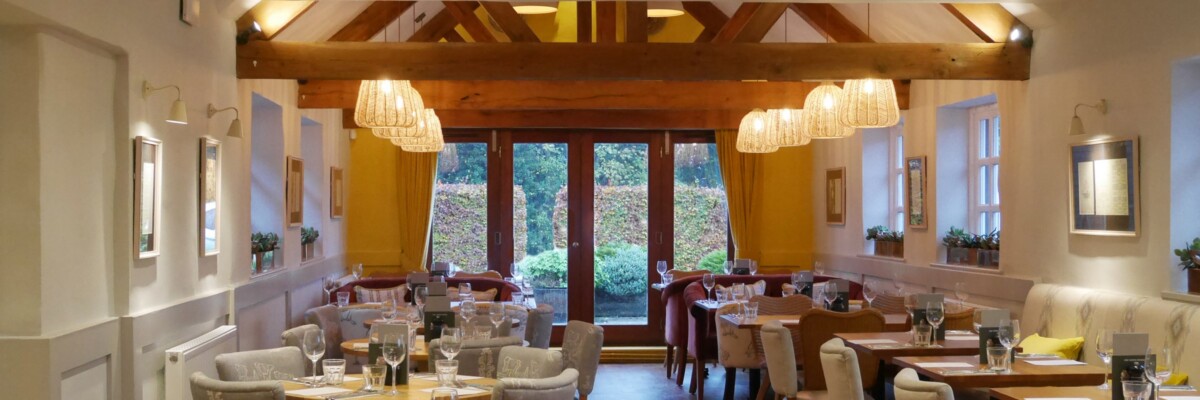 A cosy, well-lit restaurant dining area with wooden beams, modern light fittings, neatly set tables, and large windows overlooking a leafy garden.