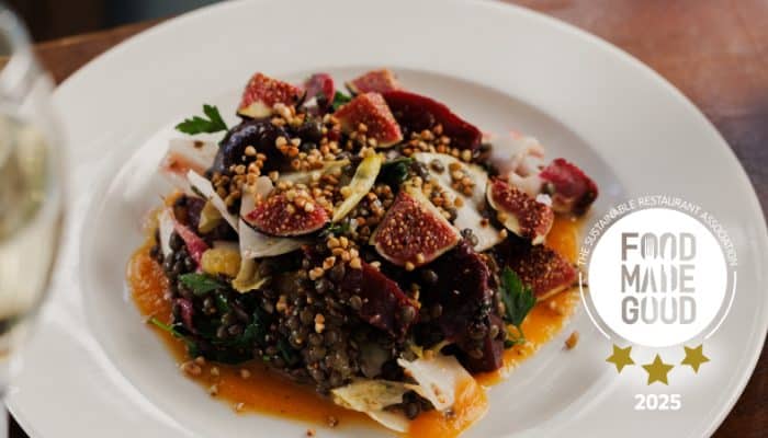 A colourful salad with lentils, sliced figs, leafy greens, and seeds sits on a white plate. An orange sauce surrounds the salad. A Food Made Good 2025 badge with three gold stars is on the plate.