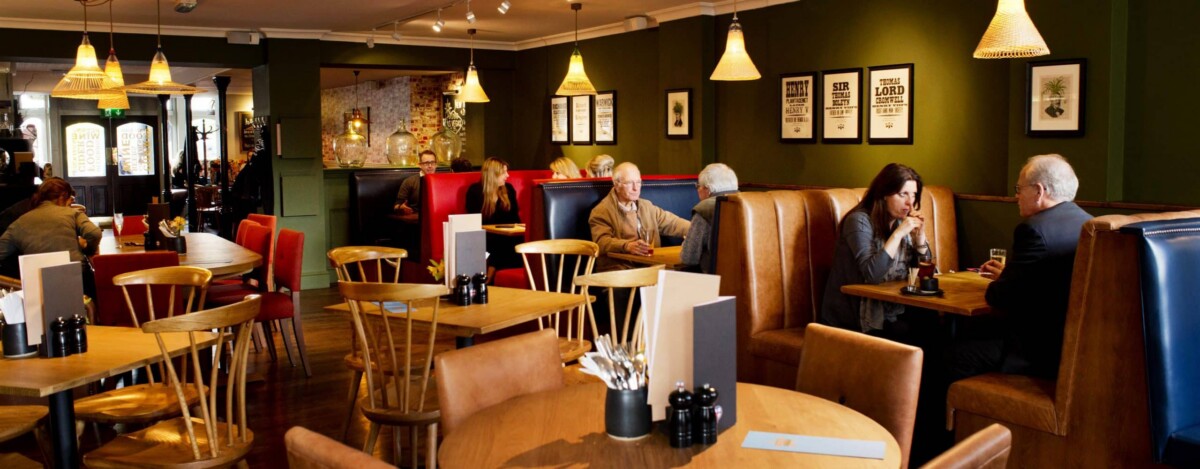 A cosy restaurant with wooden tables and chairs, green walls, pendant lights, and people sitting in booths and at tables, engaged in conversation or dining. Framed pictures decorate the walls.