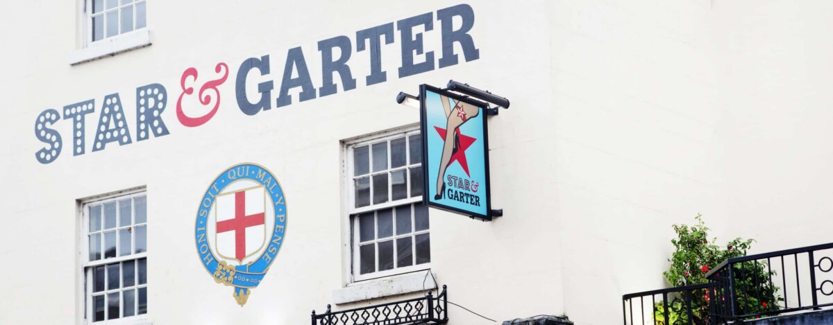 The exterior of The Star & Garter Warwickshire pub features its name painted on a cream-coloured wall above two windows, with a hanging sign showing a red star and blue background. A decorative crest and balcony with plants complete the charming scene.