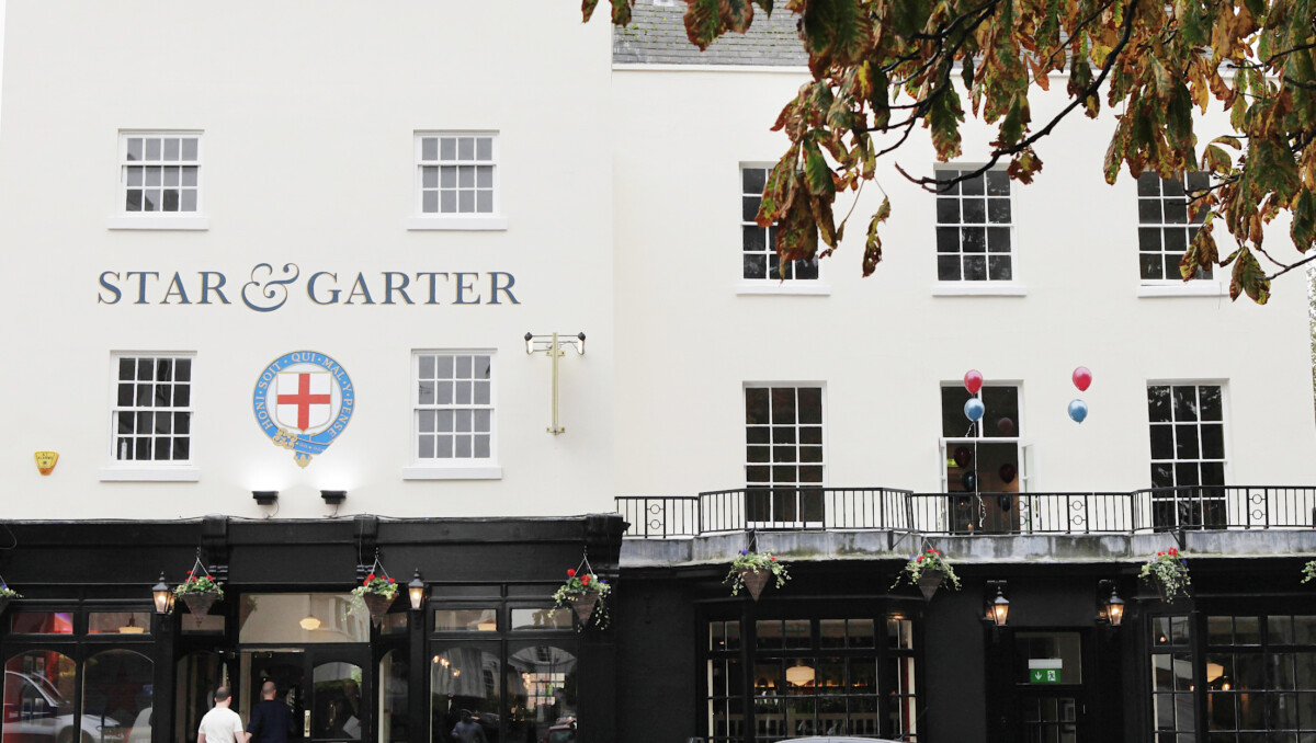 A white building with Star & Garter sign, large windows, and hanging flower baskets. Three balloons are tied to the balcony railing, and tree branches hang down from the top right corner.
