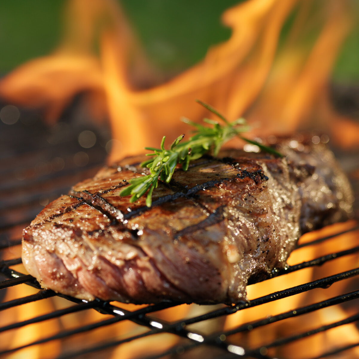 A thick, juicy steak topped with fresh rosemary is grilling over open flames on a barbecue, with char marks visible and a blurred background of orange fire.