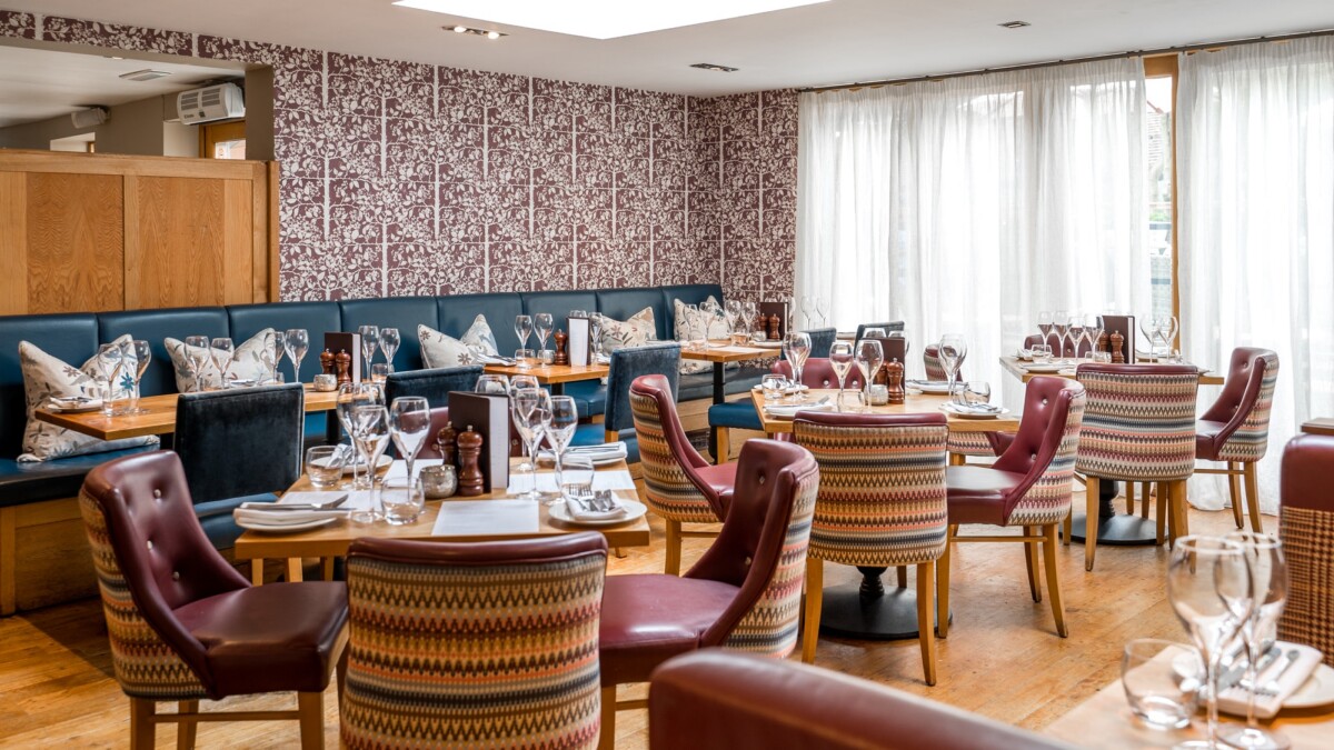 A stylish restaurant dining area with round wooden tables, red and patterned upholstered chairs, a cushioned bench along one wall, white curtains, and decorative wallpaper. Tables are set with glassware and serviettes.