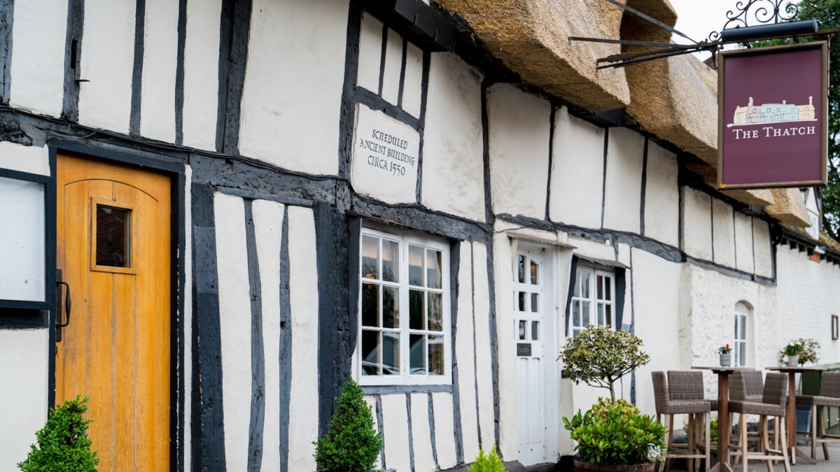 A quaint, whitewashed, half-timbered pub with a thatched roof, wooden door, and a sign reading The Thatch Oxfordshire. Outdoor tables and potted plants sit by the entrance, giving a welcoming, rustic appearance.