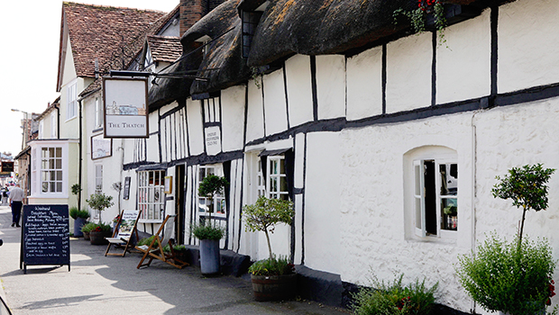 A traditional English pub with whitewashed, timber-framed walls and a thatched roof, featuring outdoor plants, a chalkboard menu, and windows with flower boxes along a quaint village street.