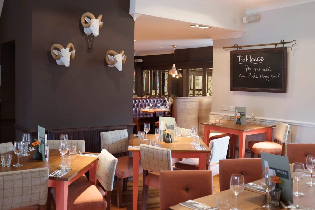 A cosy restaurant dining area with wooden tables set for meals, upholstered chairs, a blackboard wall sign reading The Fleece, and three decorative ram's heads mounted on a dark wall.