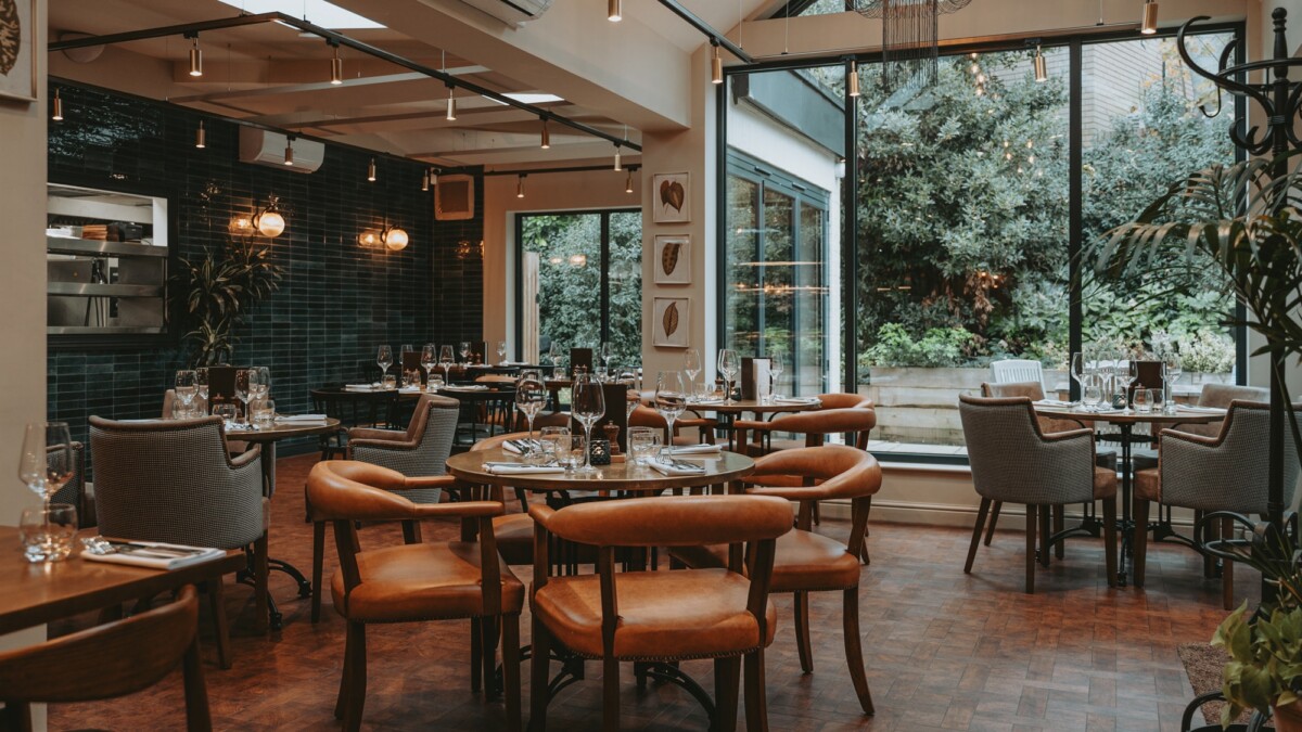 A stylish restaurant interior with wooden tables, leather chairs, and large windows overlooking a green garden. The space is warmly lit, with neatly set tables and modern décor.