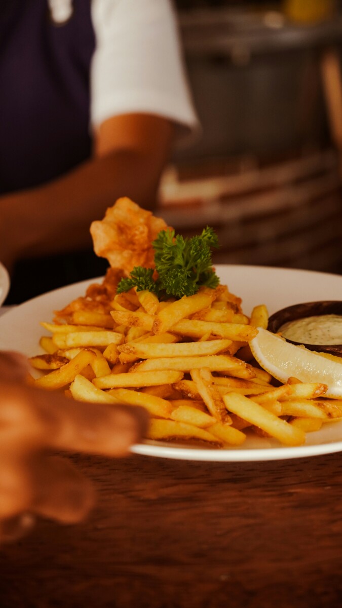 A close-up of a plate of golden chips garnished with parsley, served with a lemon wedge and a dipping sauce on the side. A person’s arm and hand are visible in the background.