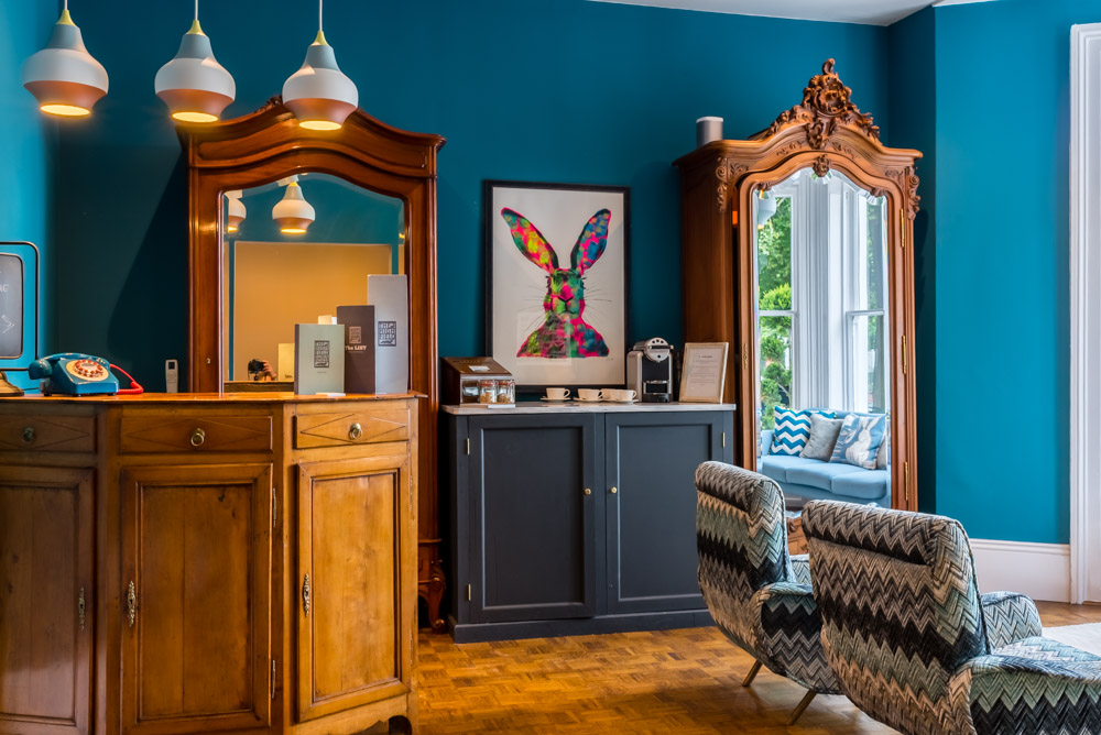 A stylish room with teal walls, vintage wooden wardrobes, modern patterned chairs, pendant lights, and a colourful rabbit artwork above a cabinet. Large windows let in natural light over a cosy window seat with blue cushions.