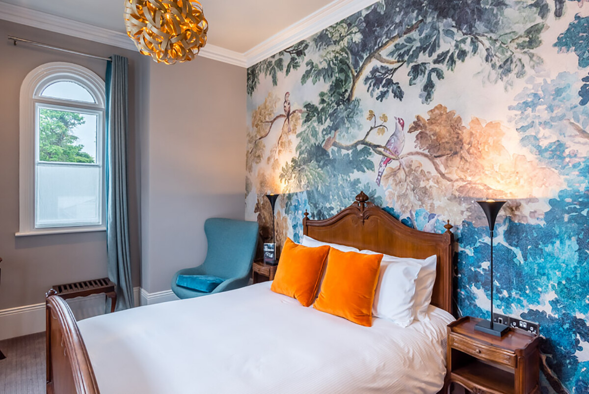 A cosy bedroom at The High Field Town House Birmingham features a wooden bed, white bedding, orange pillows, a mural of birds on tree branches, two tall lamps, a blue armchair, teal curtains on the window, and a golden chandelier.