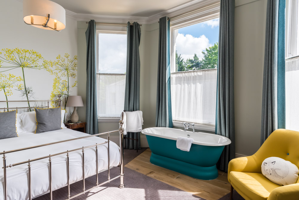 Bright bedroom at The High Field Town House Birmingham with a metal-framed bed, grey and yellow cushions, teal bath, yellow armchair, bedside table with lamp, large windows with teal curtains, and floral wall art. Sunlight streams in.
