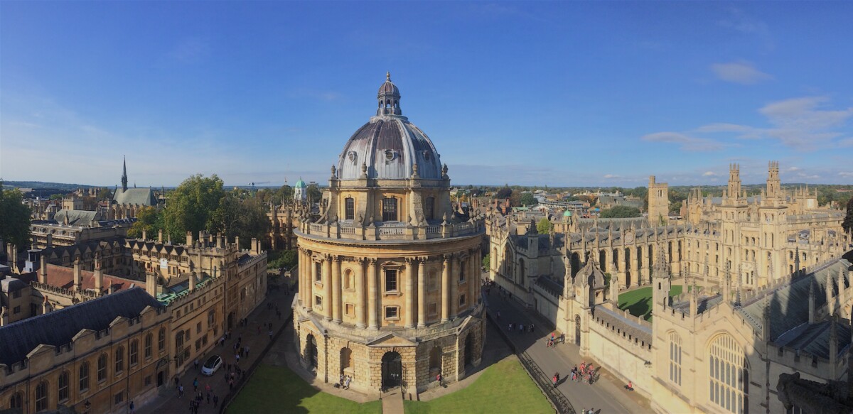 A panoramic view of the Radcliffe Camera, a large domed building, surrounded by historic Oxford University buildings under a clear blue sky. Nearby, people stroll the paths, perhaps heading towards The Bear & Ragged Staff for a classic experience.