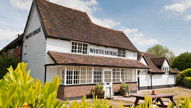 The White Horse Harpenden is a traditional English pub with white timber walls, a red tiled roof, large windows, and outdoor seating on a sunny day, all surrounded by lush greenery.