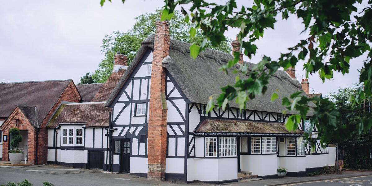 A charming, traditional English cottage with white walls, dark timber framing, a thatched roof, and red brick chimneys, set amongst greenery on a quiet street.