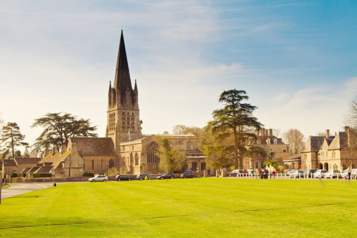 A historic stone church with a tall spire stands amid trees and old buildings, facing a wide green lawn near The Fleece Cotswold, under a blue sky with scattered clouds. Several parked cars line the road near the church.
