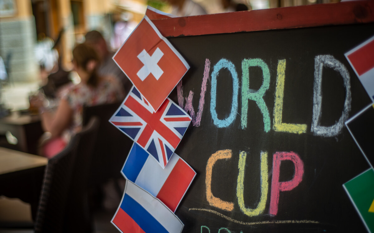 A colourful chalkboard sign reads “WORLD CUP,” decorated with small paper flags of Switzerland, the UK, France, and Italy. Blurred people are sitting in the background.